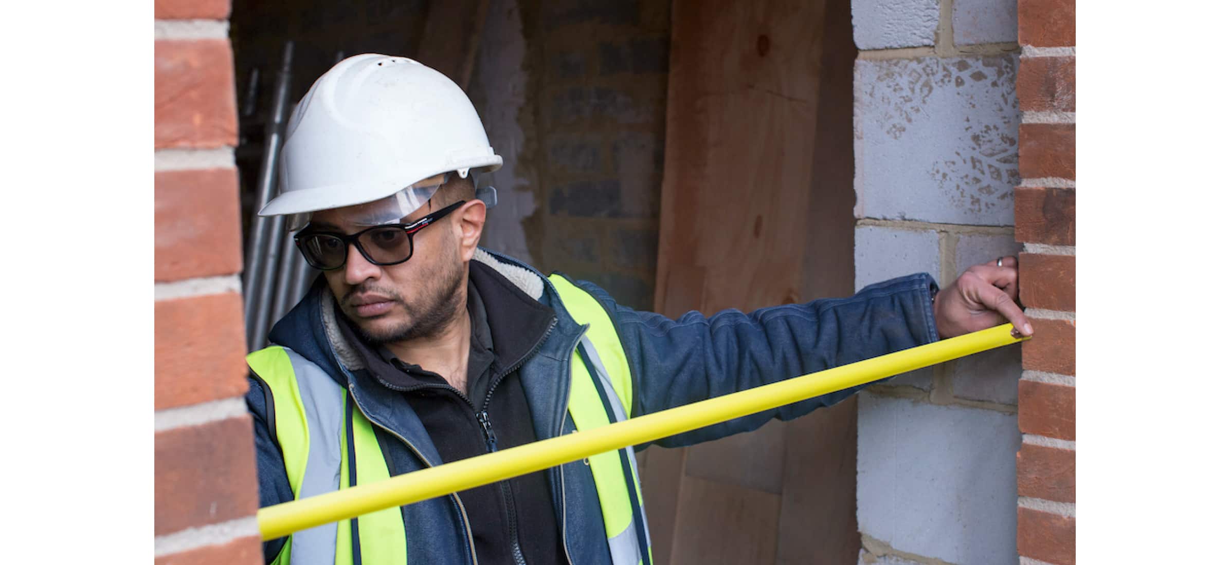 Hitesh at a construction site wearing a safety hat, measuring a doorway with a measuring tape.